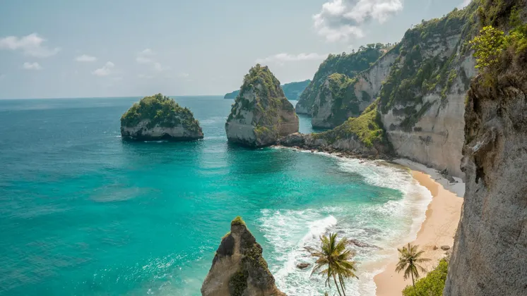 Vue plongeante d'une crique isolée avec de grandes formations rocheuses dans des eaux turquoises, une plage de sable et des falaises abruptes.