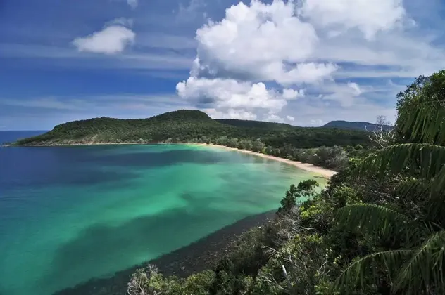 Eine Vogelperspektive auf eine tropische Bucht mit türkisfarbenem Wasser, einem Sandstrand und einem bewaldeten Hügel unter einem teilweise bewölkten Himmel.