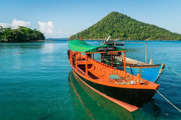 Ein farbenfrohes Longtail-Boot mit grünem Dach liegt in ruhigem, türkisfarbenem Wasser vor einem bewaldeten Hügel.