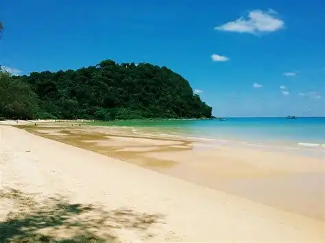 Ein breiter Sandstrand trifft auf ruhiges, türkisfarbenes Wasser neben einem üppigen, bewaldeten Hügel unter einem klaren blauen Himmel.