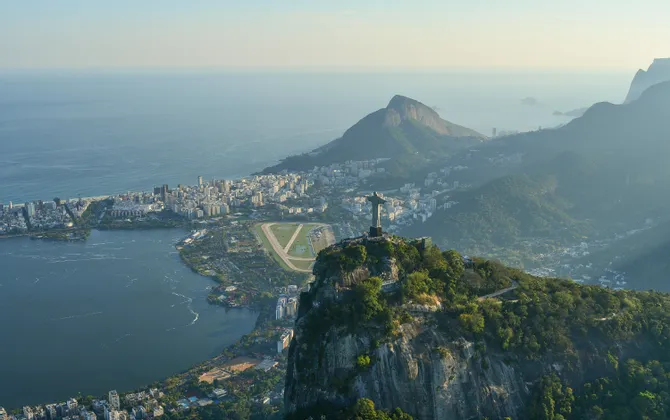 An aerial view of a large statue on a mountain peak, overlooking a coastal city with a large lagoon and the ocean in the background.