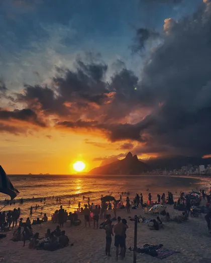 A large crowd of people on a beach watching a dramatic sunset over the ocean, with silhouetted mountains in the background.