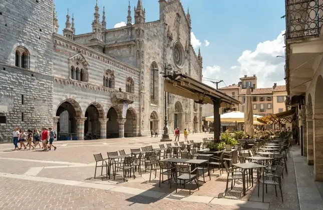 Un caffè all'aperto con tavoli vuoti si trova in una piazza soleggiata e pavimentata di fronte a una grande cattedrale in pietra a strisce.