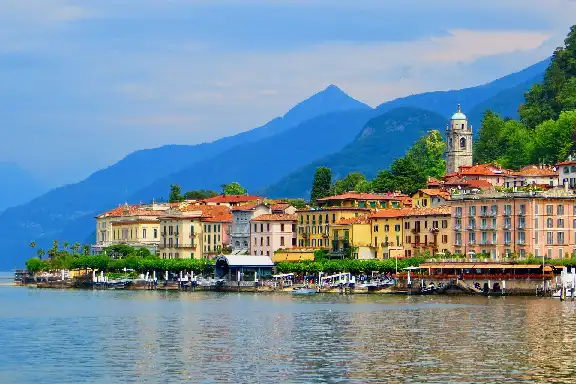 Una colorata cittadina in riva al lago con un campanile è incastonata ai piedi di grandi montagne alberate sotto un cielo azzurro.