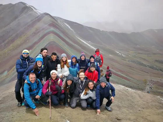 Un gruppo WeRoad in viaggio posa per una foto su una cima di montagna con formazioni rocciose colorate e stratificate sullo sfondo.