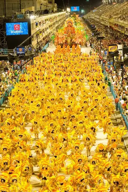 A high-angle view of a nighttime parade with hundreds of performers in bright yellow costumes, watched by crowds in large stands.