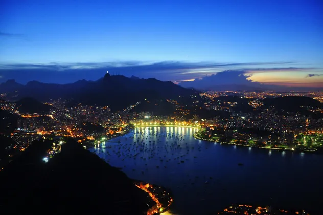 An aerial view of a coastal city illuminated at dusk, with a bay full of boats and mountains silhouetted in the background.