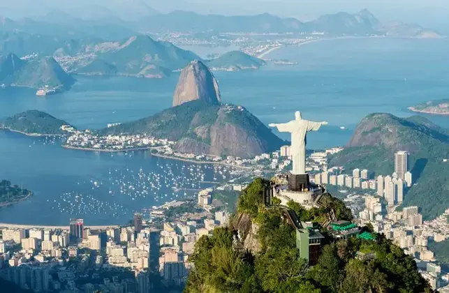 The Christ the Redeemer statue stands on a mountain peak overlooking a coastal city, Sugarloaf Mountain, and a bay filled with boats.