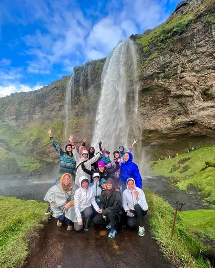 Un groupe WeRoad en imperméables sourit et pose pour une photo devant une grande cascade sur une falaise moussue.