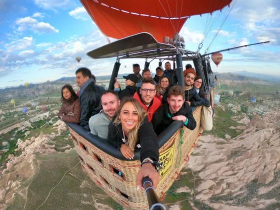 Un gruppo WeRoad si scatta un selfie all'interno di una cesta di mongolfiera che sorvola una valle rocciosa con altre mongolfiere nel cielo.