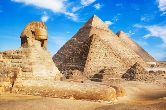 Le Grand Sphinx trône dans le désert avec les Pyramides de Gizeh en arrière-plan sous un ciel bleu.