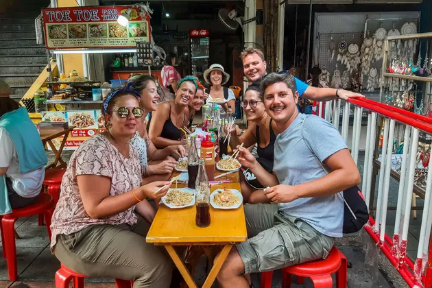 Un groupe WeRoad sourit en dégustant des nouilles avec des baguettes à un stand de street food en plein air.