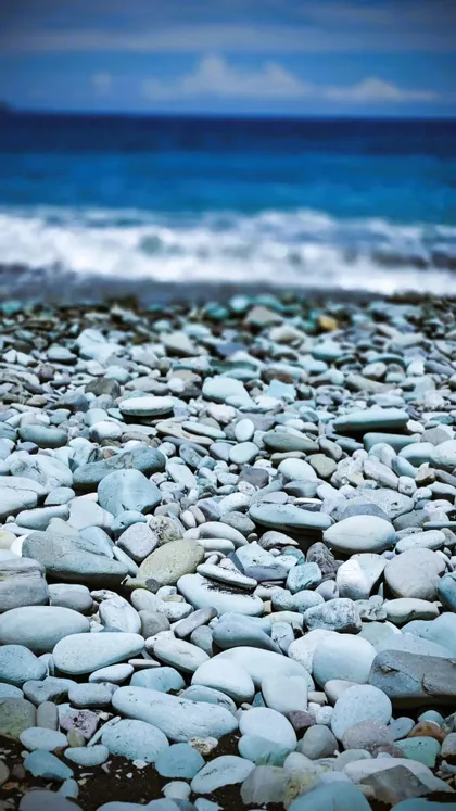 Nahaufnahme von glatten, hellblauen Kieselsteinen am Strand, mit dem tiefblauen Meer unscharf im Hintergrund.