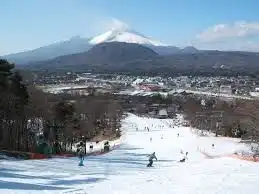 Persone che sciano e fanno snowboard su un'ampia pista con una città e un grande vulcano innevato sullo sfondo.
