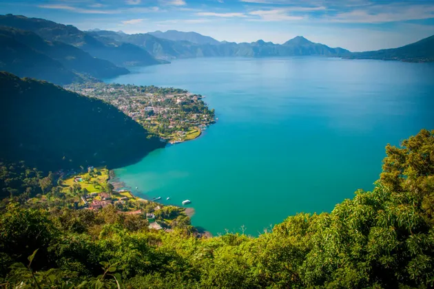 Panoramablick auf einen großen türkisfarbenen See, umgeben von grünen Bergen und einer Stadt am Ufer.
