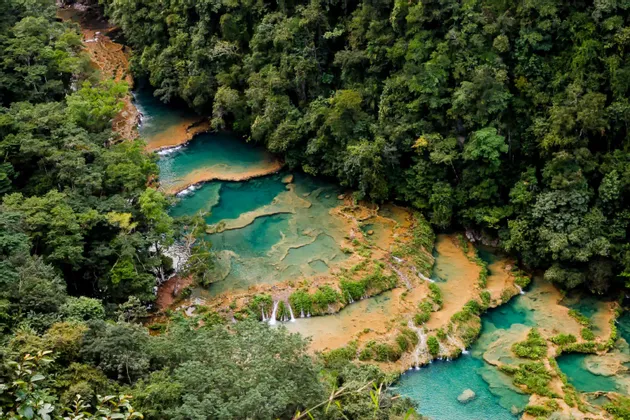 Eine Luftaufnahme eines Flusses mit türkisfarbenen, terrassenförmigen Becken und kleinen Wasserfällen, die durch einen dichten, grünen Dschungel fließen.