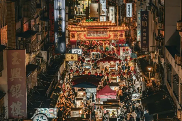 Eine Vogelperspektive auf einen belebten asiatischen Nachtmarkt, auf dem Menschen zwischen Ständen unter beleuchteten Schildern und roten Laternen umhergehen.