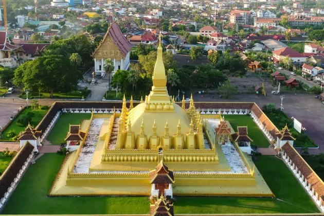 Eine Luftaufnahme einer großen, goldenen Stupa mit mehreren Türmen, umschlossen von einem ummauerten Hof, mit einer Stadt im Hintergrund.