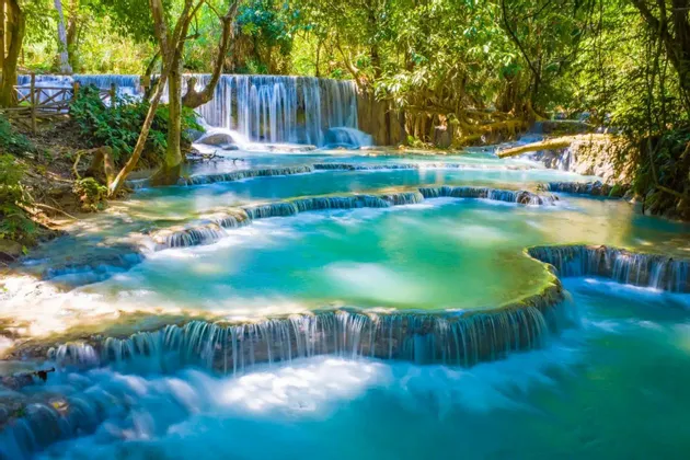 Ein breiter, mehrstufiger Wasserfall fließt in mehrere türkisfarbene Becken in einem sonnendurchfluteten grünen Wald.
