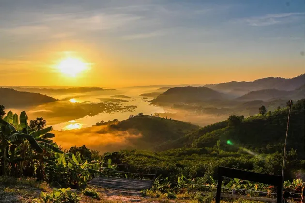 Goldener Sonnenaufgang über einem breiten Fluss, der sich durch neblige, dschungelbedeckte Berge schlängelt, gesehen von einem hölzernen Aussichtspunkt im Vordergrund.