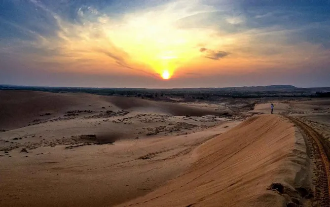 Una persona si erge sulla cresta di una grande duna di sabbia, osservando il tramonto su un vasto paesaggio desertico.