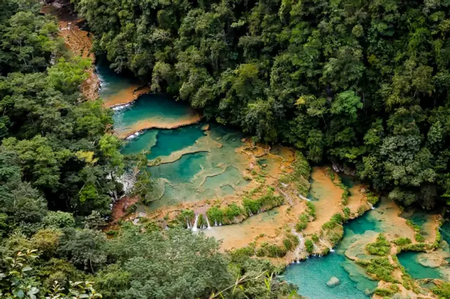 Eine Luftaufnahme von türkisfarbenem Flusswasser, das über terrassierte Becken und Wasserfälle fließt, umgeben von dichtem grünen Dschungel.