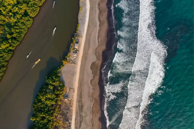 Eine Luftaufnahme eines Flusses mit Booten neben einem Sandstrand, der an den türkisfarbenen Ozean mit weißen Wellen grenzt.