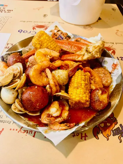 A close-up of a metal tray with a seafood boil of shrimp, crab legs, clams, corn, and potatoes covered in a red sauce.