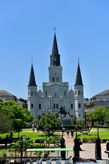 A large white cathedral with three steeples stands behind a green park containing an equestrian statue and people walking.