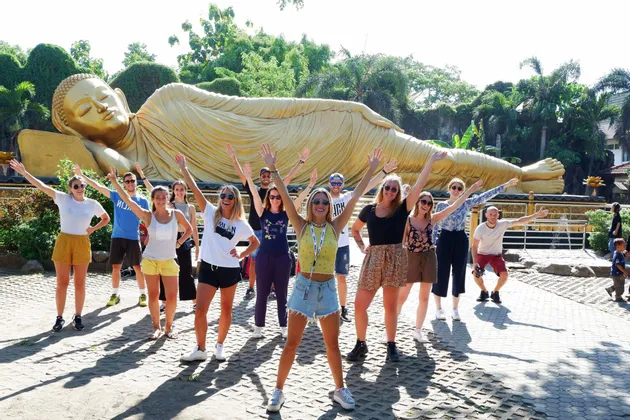 Eine WeRoad-Gruppe posiert mit erhobenen Armen vor einer riesigen, goldenen, liegenden Buddha-Statue.