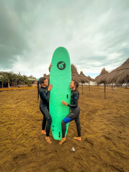 Due donne in mute nere si sorridono, tenendo una tavola da surf verde in verticale su una spiaggia sabbiosa sotto un cielo nuvoloso.