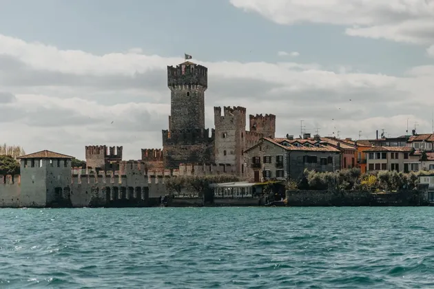 A medieval stone castle with fortified walls and towers stands on the shore of a body of water under a partly cloudy sky.