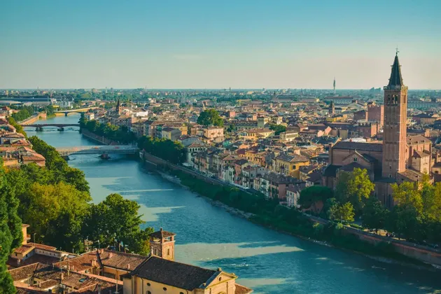 An aerial view of a historic city with a wide river, multiple bridges, terracotta roofs, and a prominent bell tower.