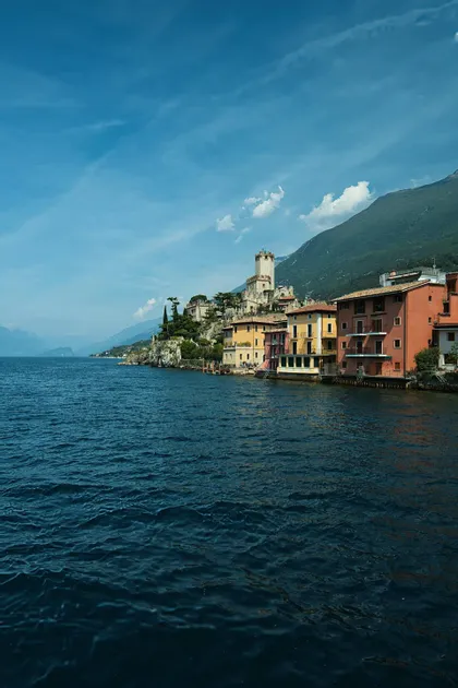 A lakeside village with colorful houses and a castle on a cliff, seen from the water with a mountain behind.