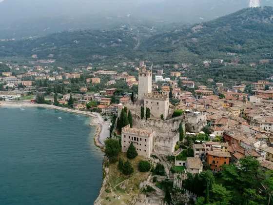 An aerial view of a stone castle on a cliff overlooking a lakeside town, with green mountains in the background.