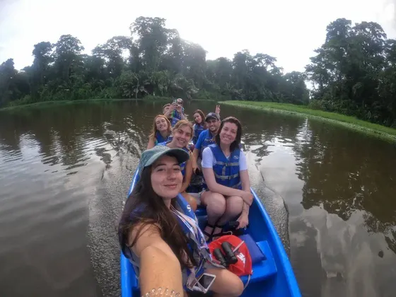 Eine WeRoad-Gruppe, ausgestattet mit Schwimmwesten, macht ein Gruppenselfie auf einem blauen Boot, das einen von dichtem Dschungel gesäumten Fluss befährt.