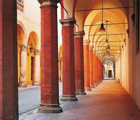 Un lungo portico con una fila di colonne ottagonali in mattoni rossi, un soffitto a volta e un'illuminazione calda da lampade a sospensione.