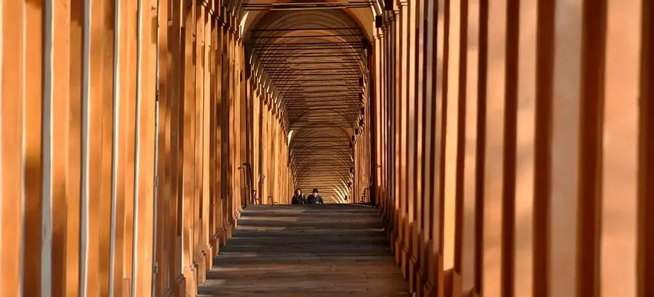 Due persone sono sedute su dei gradini in lontananza, lungo un lungo portico ad arco con colonne di terracotta e ombre che si proiettano dalla luce del sole.
