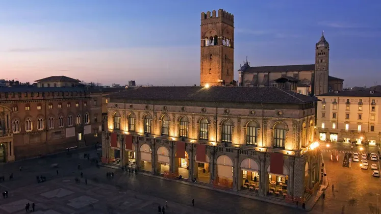 Una vista dall'alto di una piazza storica cittadina al crepuscolo, con edifici illuminati e un'imponente torre in mattoni