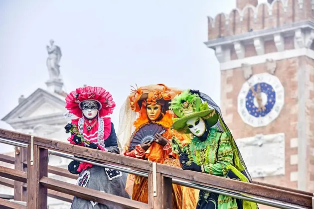 Trois personnes en costumes et masques de carnaval vénitiens colorés se tiennent sur un pont devant une tour de l'horloge.