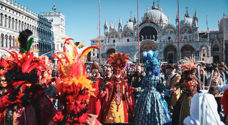 Une foule de personnes portant des masques de carnaval élaborés et colorés et des costumes à plumes célèbrent sur une place historique.