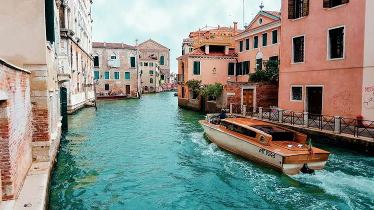 Un bateau à moteur en bois navigue sur un canal aux eaux turquoise, bordé de vieux bâtiments colorés dans une ville historique.