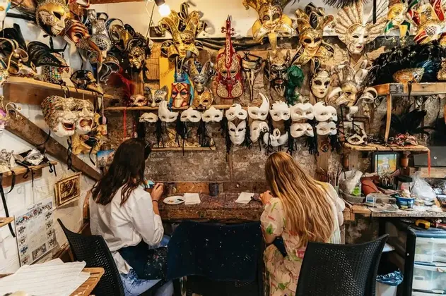 Deux femmes participant à un voyage de groupe WeRoad peignent des masques à une table dans un atelier rempli de masques vénitiens.