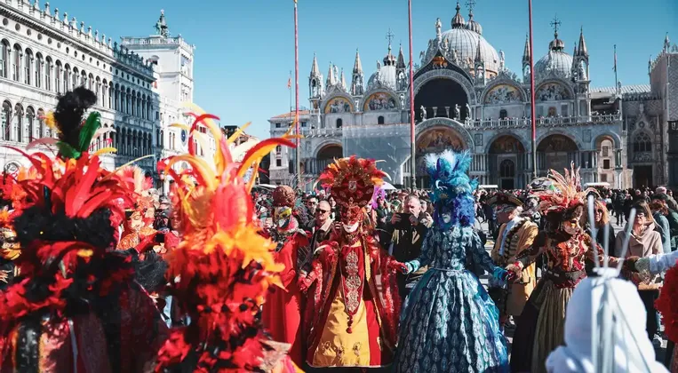 Une foule en costumes colorés et masques à plumes célèbre un carnaval sur une place historique.