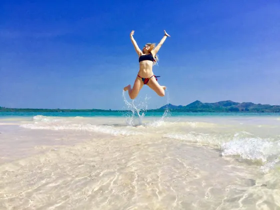 Una donna in bikini salta fuori dall'acqua bassa e cristallina di una spiaggia tropicale, con le braccia alzate verso il cielo blu.