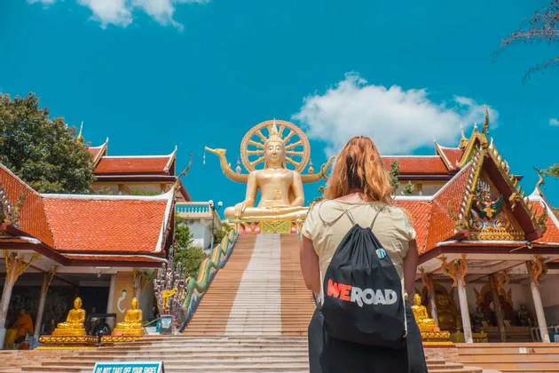 Una mujer con una mochila WeRoad mira hacia una larga escalera que conduce a una gran estatua de Buda dorado en un complejo de templos.