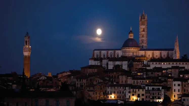 Una città storica con una cattedrale e torri illuminate, sotto un cielo notturno buio con una luna piena splendente.