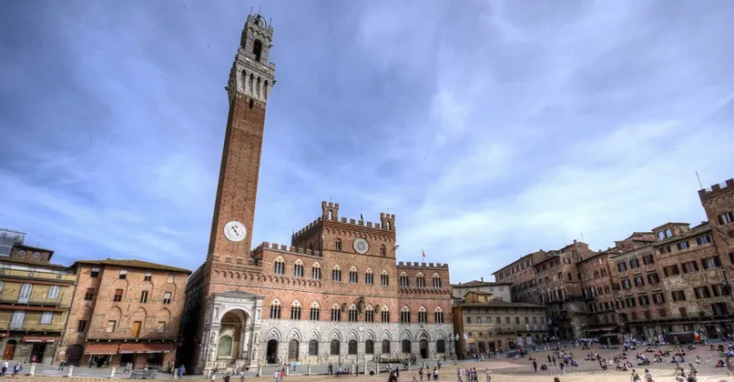 Un'alta torre dell'orologio in mattoni e un edificio storico adiacente dominano una piazza pubblica affollata sotto un cielo azzurro.