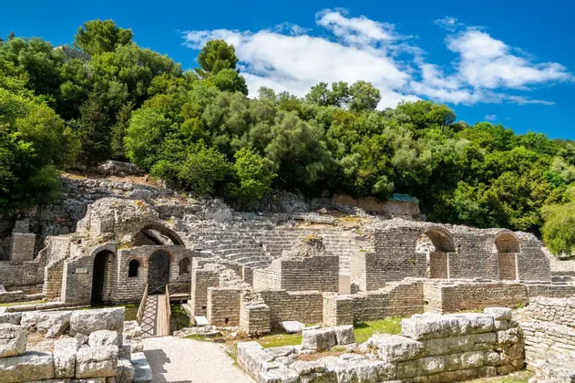 Alte Steinruinen eines Amphitheaters, eingebettet in einen von grünen Bäumen bewachsenen Hügel unter blauem Himmel.