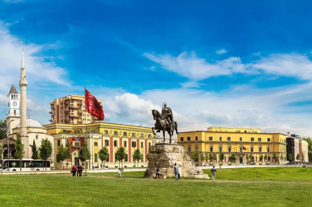 Eine bronzene Reiterstatue auf einem Steinsockel auf einem begrünten Platz, mit einer großen albanischen Flagge und historischen Gebäuden im Hintergrund.